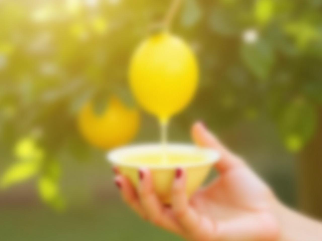 A close-up of a womans hand squeezing fresh lemon juice into a small bowl, with a blurred background of a lemon tree and a sunny garden.